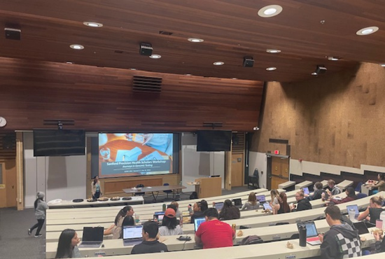 A lecture style classroom depicting the back of student heads, viewing a PowerPoint screen