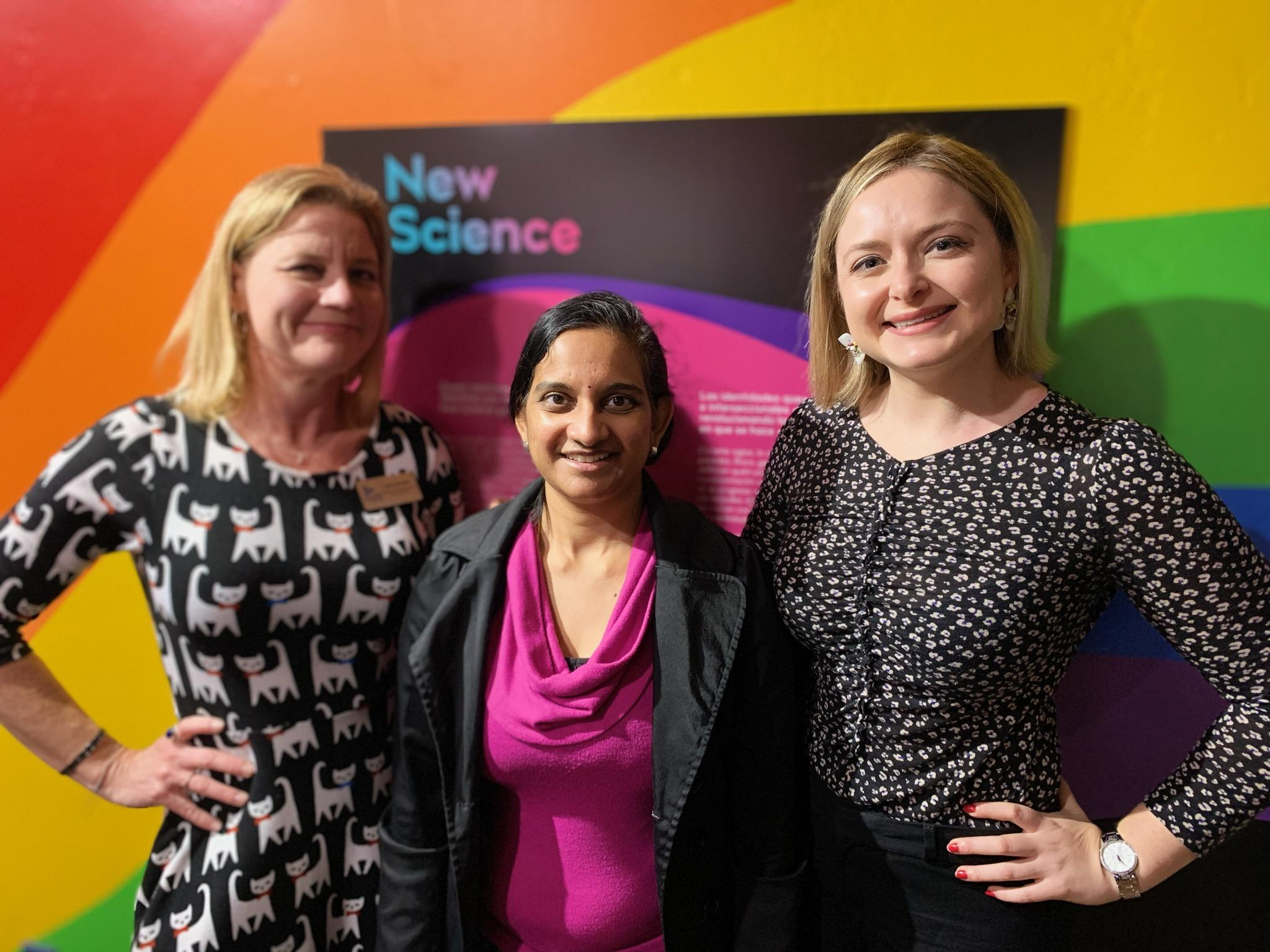 Center member Ramya Rajagopalan and Caryn Rubanovich stand next to a member of The Fleet Science Center in front of a rainbow backdrop at The Fleet Science Center.