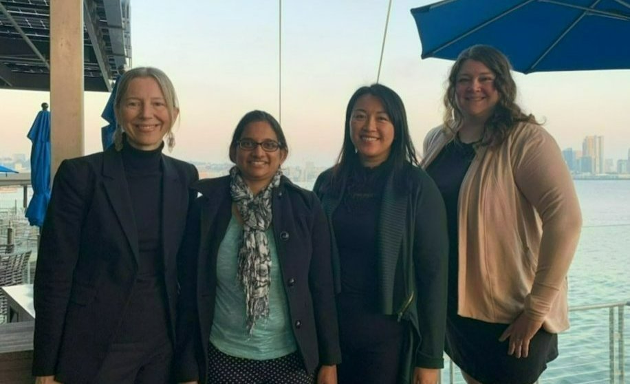 Center Director Cinnamon Bloss stands in a line next to Associate Director Ramya Rajagopalan, former center member Cynthia Triplett, and Associate Director Taylor Berninger. The four women are in a line with the background of San Diego Bay and the City of San Diego skyline.