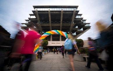 Blurred images of students walk toward a rainbow colored balloon arch in front of the Geisel Library on the UC San Diego Campus